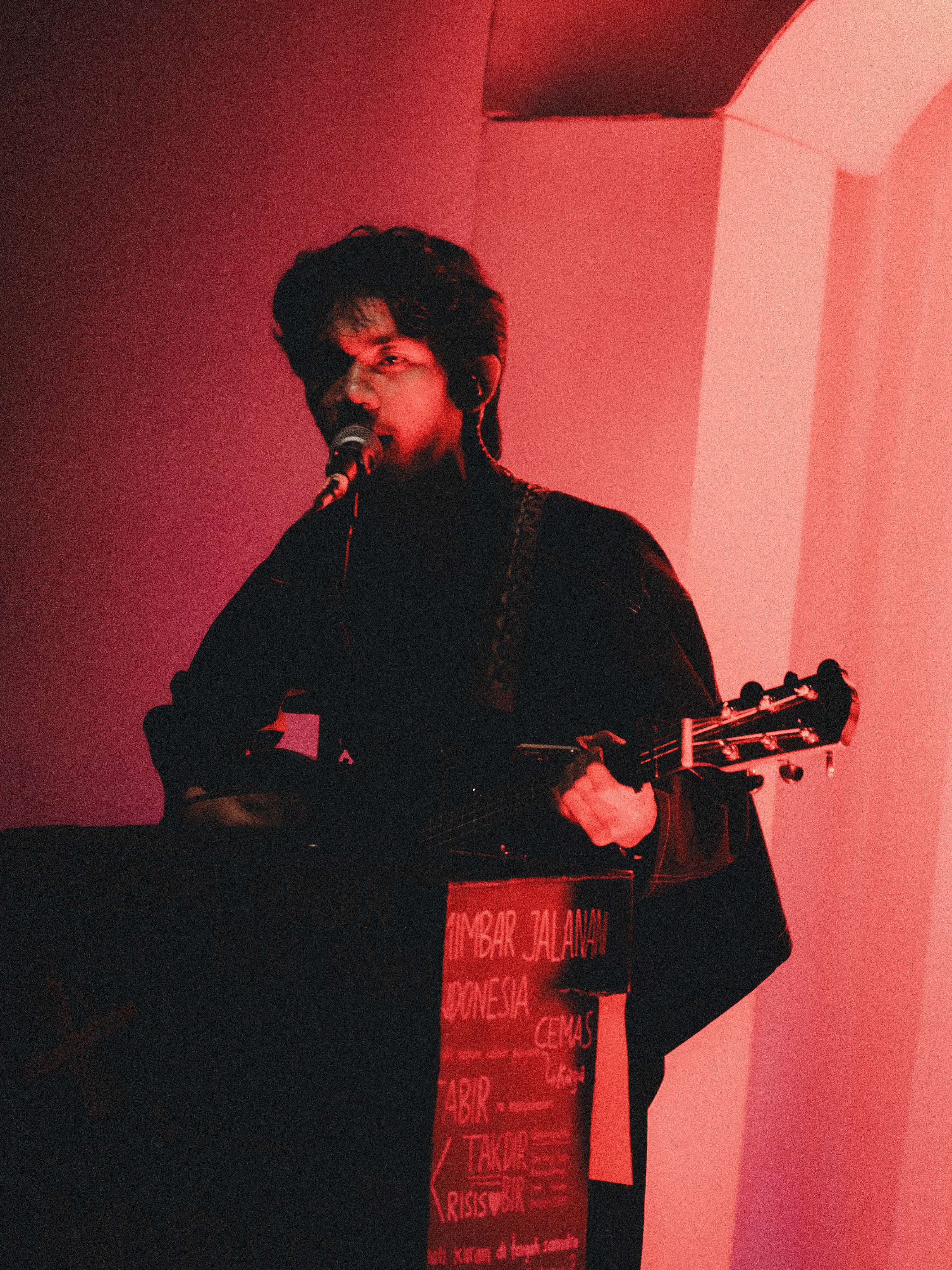 Aldy Amis bernyanyi memegang gitar dengan pencahayaan merah dramatis